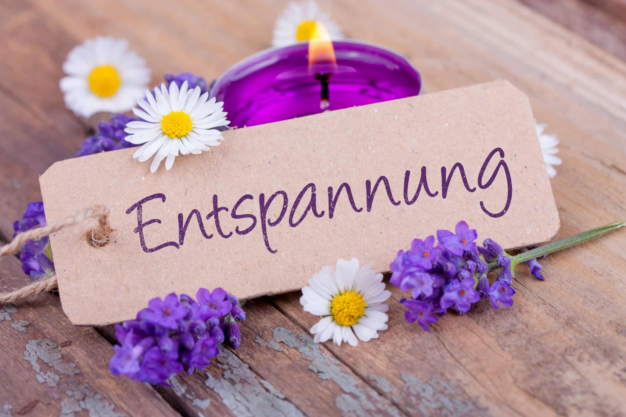 Ein „Entspannung“-Schild, umgeben von Gänseblümchen, lila Blumen und einer brennenden lila Kerze auf einer Holzoberfläche.