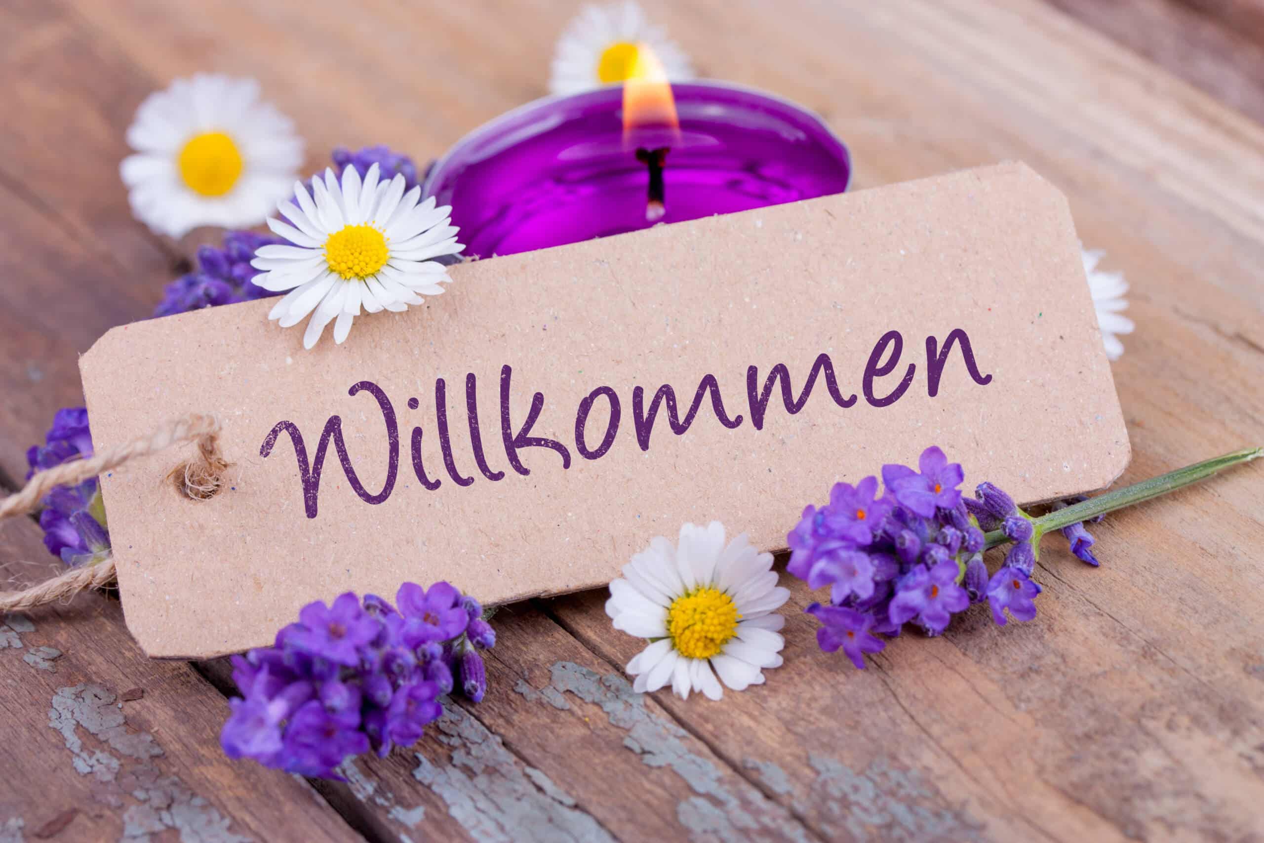 Rustikales „Willkommen“-Schild, umgeben von lila und weißen Blumen auf einer Holzoberfläche mit einer lila Kerze.
