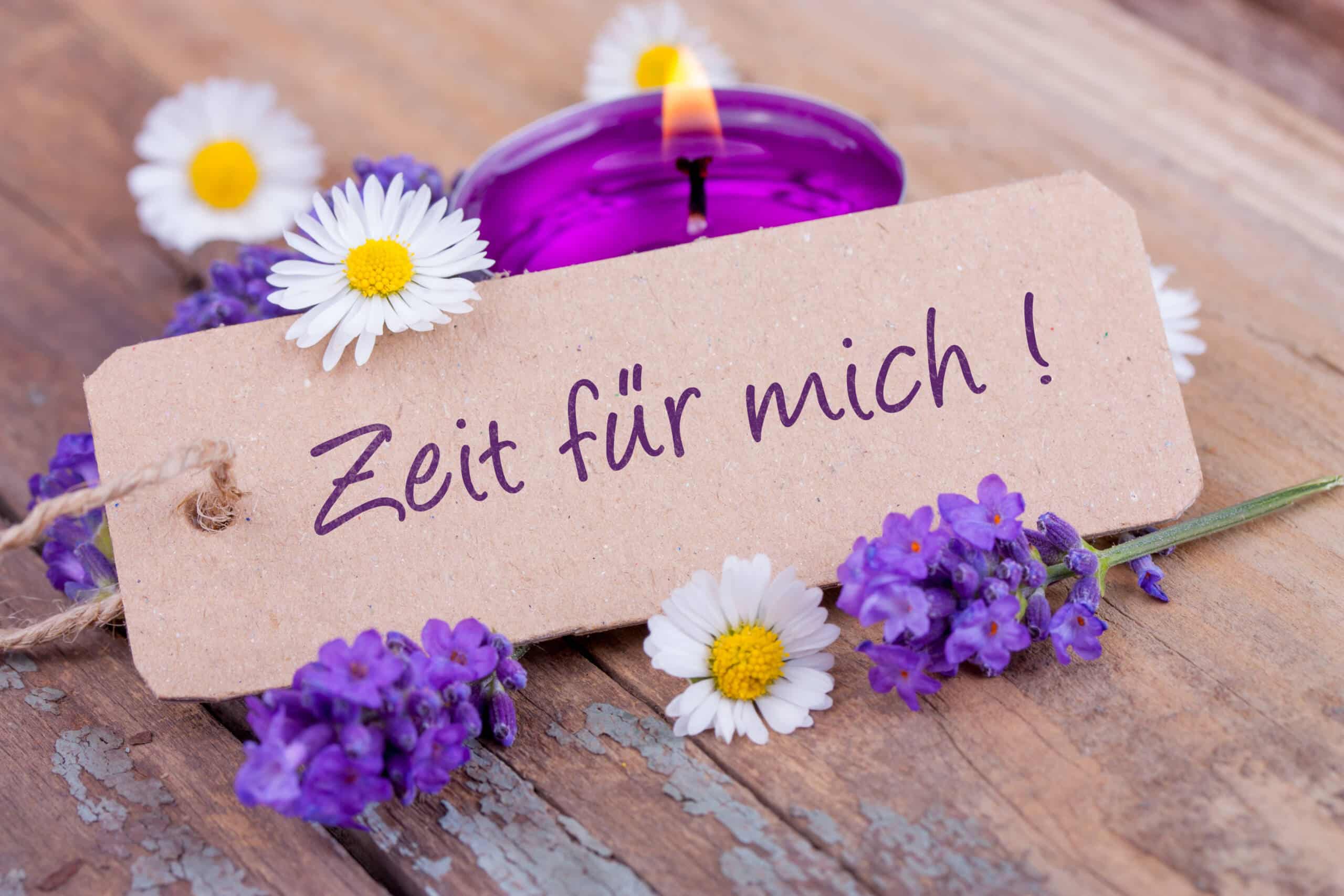 Schild mit „Zeit für mich!“ neben Blumen und einer lila Kerze auf einer Holzoberfläche.