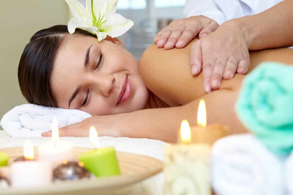 Eine Frau genießt eine Aromamassage, umgeben von Kerzen und Handtüchern, mit einer Blume im Haar und einem heiteren Gesichtsausdruck, der von purer Antistress-Glückseligkeit zeugt.